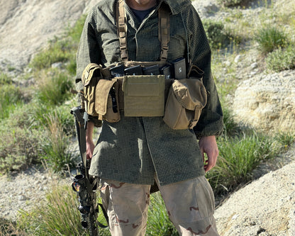 A person is standing outdoors wearing tactical gear, including a green camouflaged jacket and beige camouflaged pants. They are also equipped with the Six Gun Surplus Unissued US Marine Corps TAP Chest Rig Kit, featuring various MOLLE pouches. Holding a rifle, they stand against a backdrop of rocky terrain and grass.