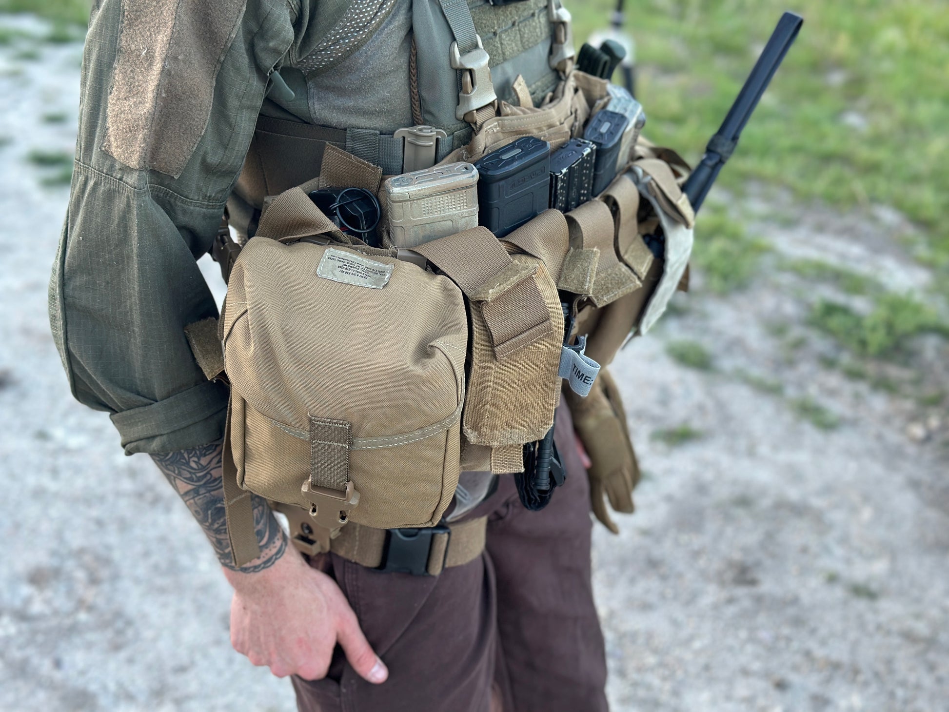 A person wearing tactical gear, including an Unissued US Marine Corps TAP Chest Rig Kit from Six Gun Surplus with multiple MOLLE pouches and magazine clips, stands outdoors on a gravel path with grass in the background. Their tattooed arm holds a black item, possibly a radio antenna. The Tactical Assault Panel integrates seamlessly into their low-profile platform.