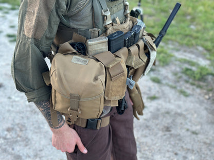 A person wearing tactical gear, including an Unissued US Marine Corps TAP Chest Rig Kit from Six Gun Surplus with multiple MOLLE pouches and magazine clips, stands outdoors on a gravel path with grass in the background. Their tattooed arm holds a black item, possibly a radio antenna. The Tactical Assault Panel integrates seamlessly into their low-profile platform.