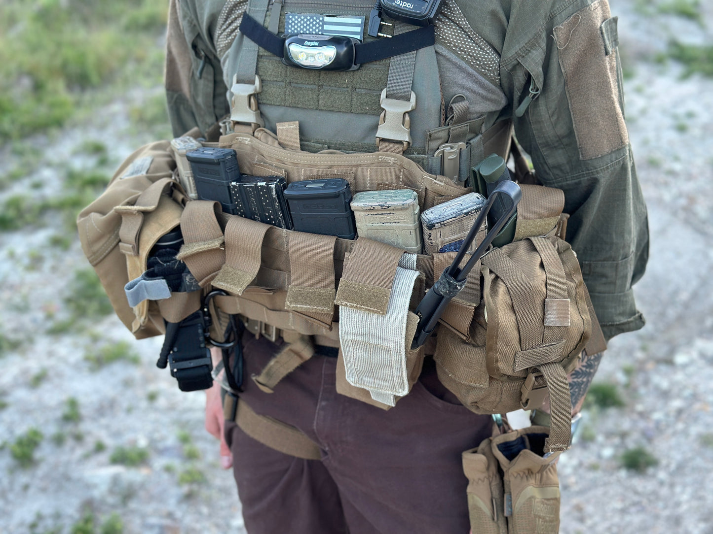 A person equipped with the Six Gun Surplus Unissued US Marine Corps TAP Chest Rig Kit, featuring multiple MOLLE pouches, a radio, and various attached equipment. The individual is dressed in a green combat shirt and brown pants while standing outdoors on rocky terrain.