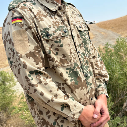 A person stands in a Six Gun Surplus German Bundeswehr Tropentarn Field Shirt, with the flag on the shoulder, blending seamlessly into the arid environment. The background reveals a dry, grassy landscape with rolling hills and scattered shrubs.