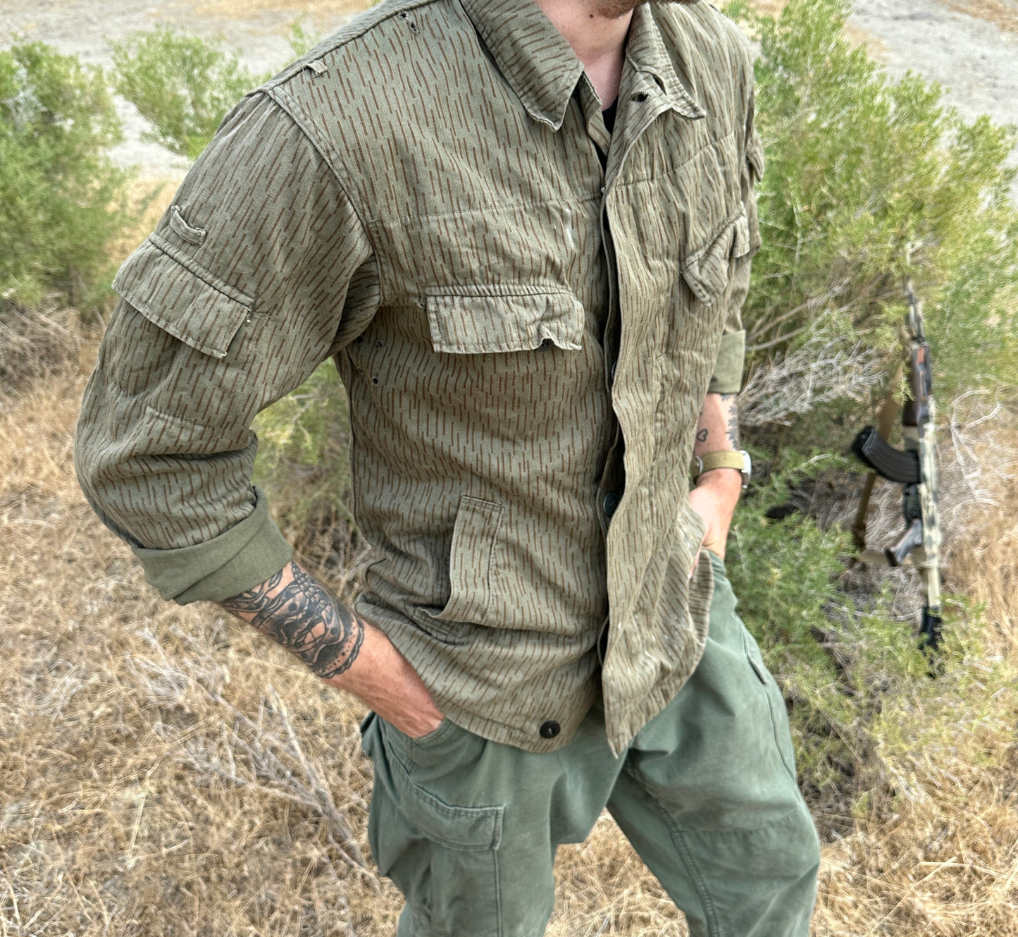 A person wearing a Six Gun Surplus East German Strichtarn Field Shirt with rolled-up sleeves and green cargo pants stands outdoors in a grassy area. The person's left arm is tattooed, and a rifle is partially visible propped against shrubs in the background, blending seamlessly with the East German Strichtarn camouflage pattern.