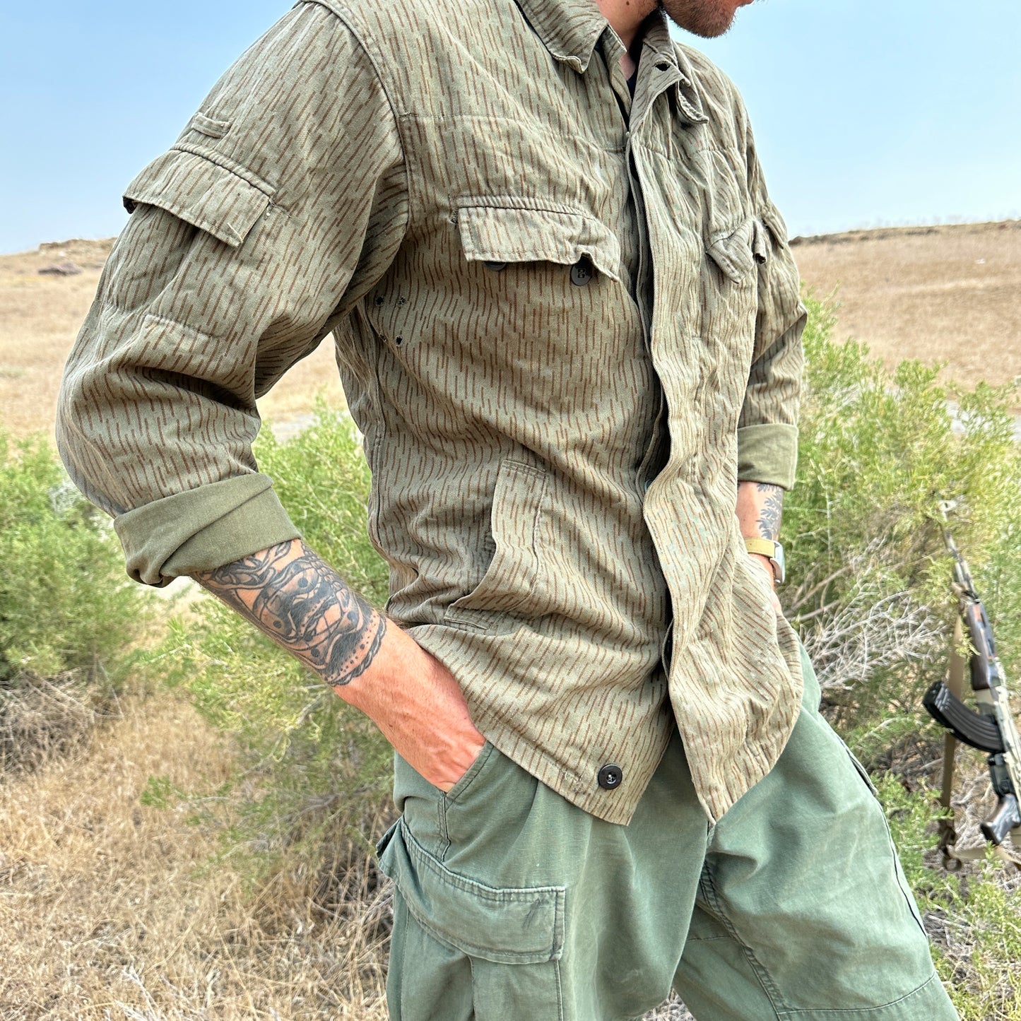 A person standing outdoors in a field, wearing the Six Gun Surplus East German Strichtarn Field Shirt—an olive-green, moisture-wicking button-up shirt with vertical stripes—and matching cargo pants in the same pattern. They have a visible arm tattoo and are holding a rifle while half-tucked in the background.