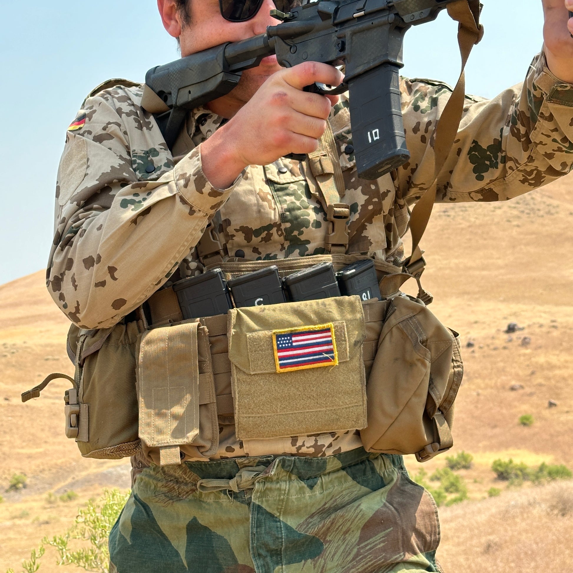 A soldier clad in a German Bundeswehr Tropentarn Field Shirt from Six Gun Surplus holds a rifle and adjusts magazine pouches on a tactical vest adorned with an American flag patch. The backdrop reveals dry, hilly terrain under a clear sky characteristic of arid environments.