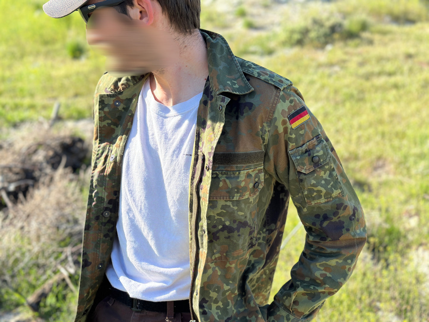 A person with a blurred face stands outdoors on grass, wearing sunglasses, a cap, a white T-shirt, and a Six Gun Surplus German Bundeswehr Flecktarn Field Shirt adorned with a flag patch on the sleeve—demonstrating practical field gear from the German Armed Forces.