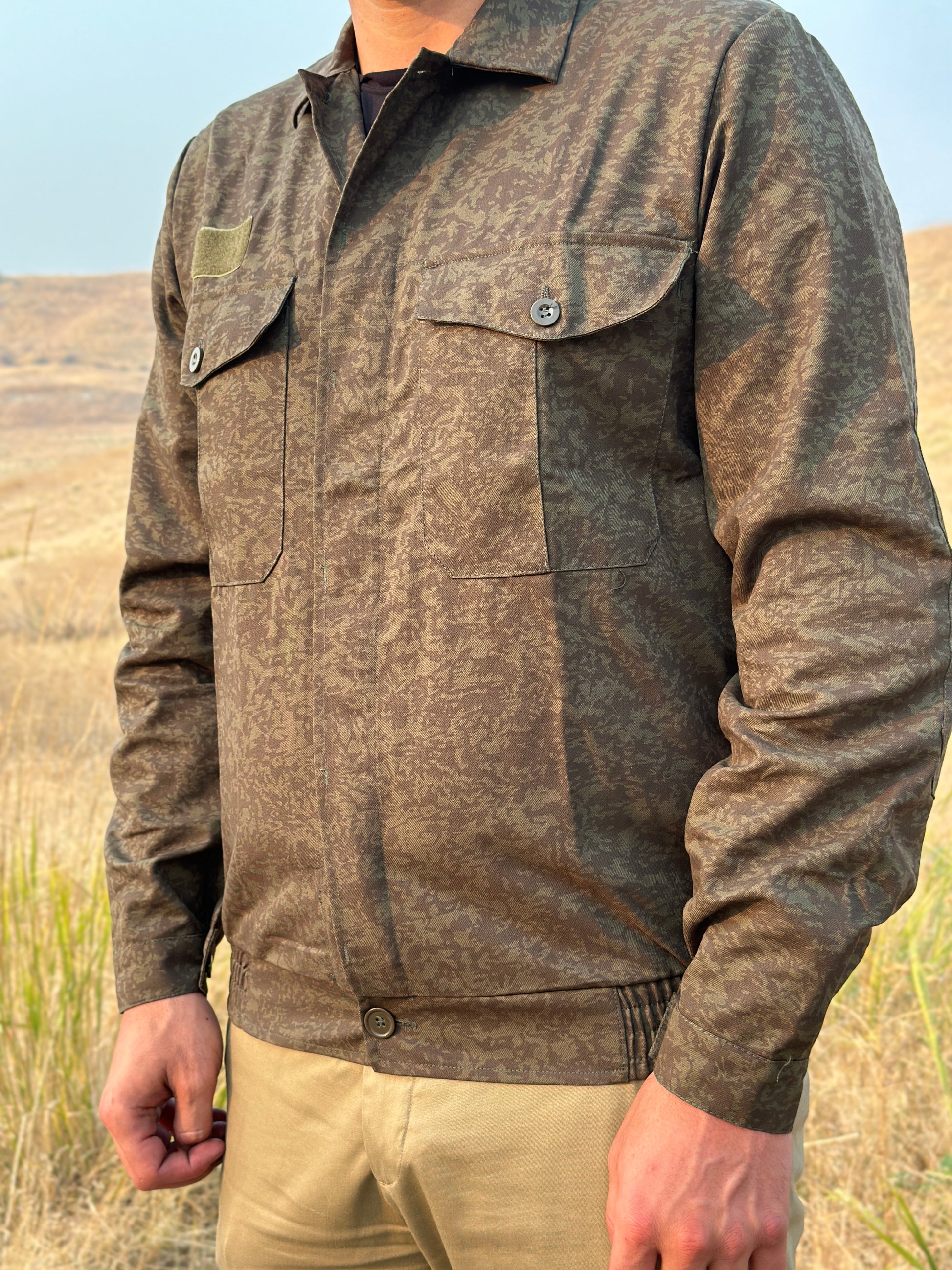 A person wearing a Czech Dark Leaf Camo Field Shirt from Six Gun Surplus, featuring reinforced elbows and button-close cargo chest pockets, stands in a dry, grassy field with hills in the background. The focus is on the shirt's details. The person's head is out of the frame.