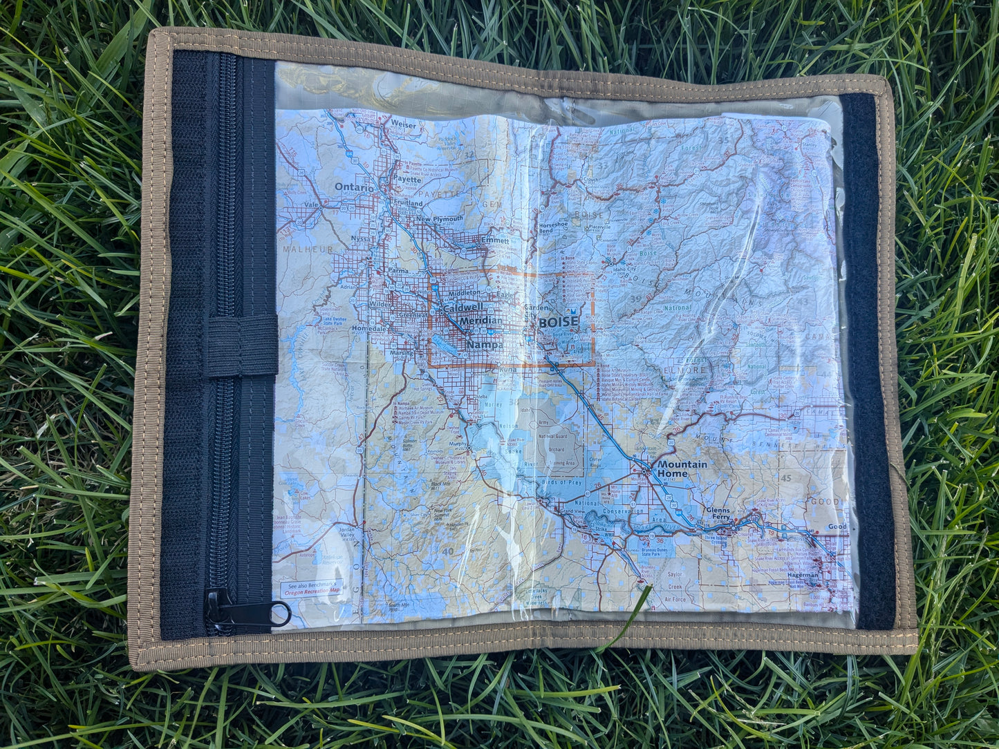 An open Map Case "RECCE pouch" from Six Gun Surplus lies on green grass, displaying a detailed topographic map of southwest Idaho, including Boise. The map is protected by a clear plastic cover and is enclosed in a brown fabric case with a black zipper.