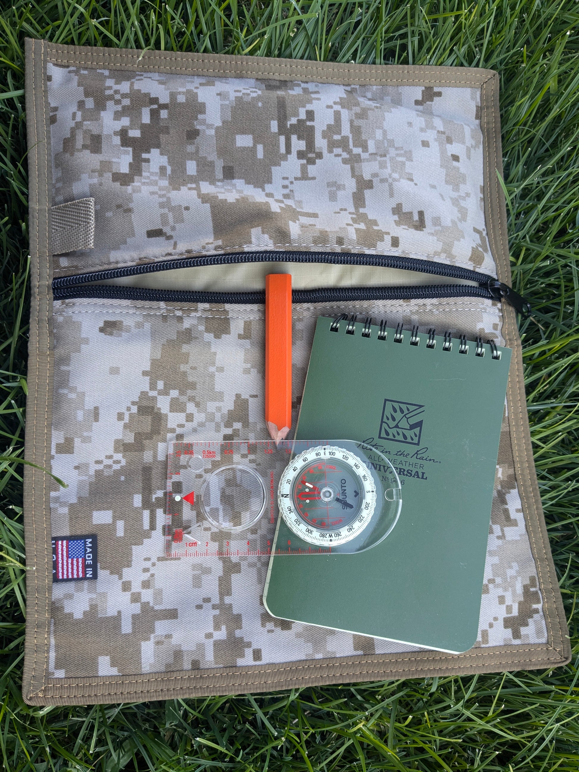 A notebook, a transparent compass, and an orange pen lie on the Six Gun Surplus Map Case "RECCE pouch" with a zipper. The items are arranged against a grassy background. The notebook has a green cover with writing on it, and the pouch features a small American flag patch.