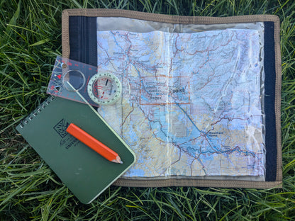 A Six Gun Surplus Map Case "RECCE pouch" is placed on green grass, featuring a detailed map of Boise, a transparent compass, a small protractor, and a green Rite in the Rain notebook accompanied by an orange pencil.