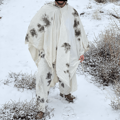 A person wearing a Six Gun Surplus German Snow Camo Overwhite (Scheentarn) poncho stands in a snowy outdoor setting. The German Army-inspired outfit, with black and gray splotches on a white background, blends seamlessly with the terrain. Sparse winter vegetation surrounds them, and footprints are visible in the snow.